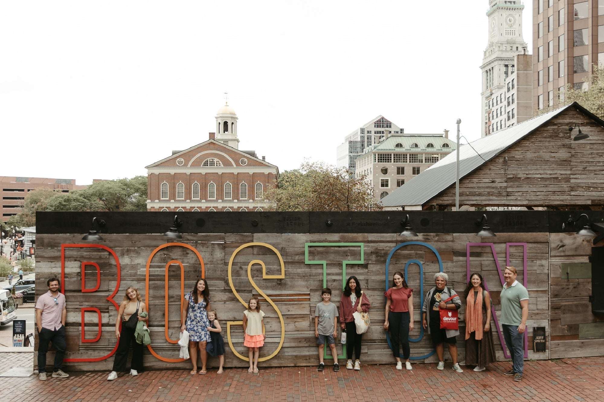 A group of people stands in front of a wooden wall with large colorful letters spelling "BOSTON," with historic buildings visible in the background.