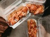 Three clear plastic containers each hold a lobster roll with chunks of lobster meat on a split bun. The containers are being held up, two with closed lids and one with the lid open.