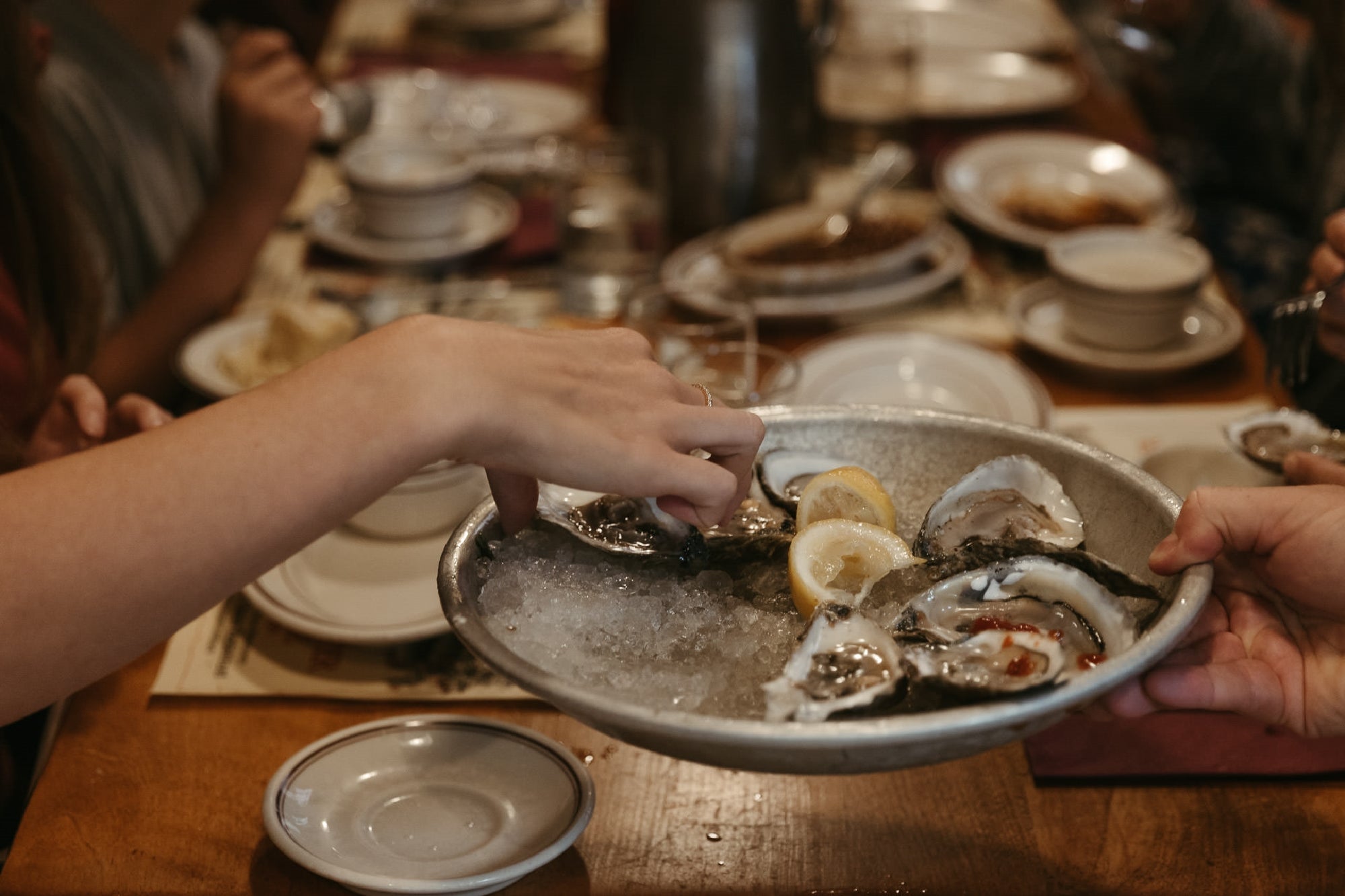 A person takes a raw oyster from a metal plate filled with oysters, lemon slices, and ice at a dining table set with dishes.