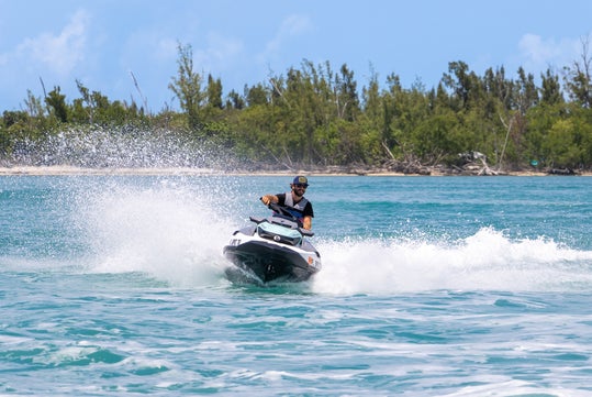 A person rides a jet ski, creating a spray of water, with a backdrop of turquoise water and a tree-lined shore.