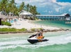 Two people ride a jet ski near a tropical beach with palm trees, loungers, and white sand. A pier extends into the blue-green water under a partly cloudy sky.
