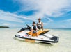 Two people wearing life jackets sit on a jet ski in shallow, clear water under a bright blue sky.