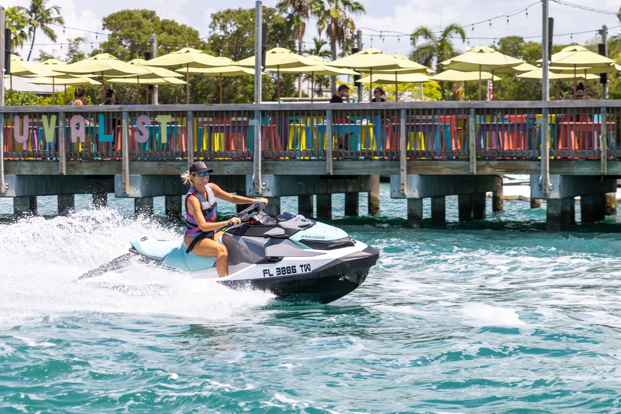 A person rides a jet ski on a sunny day near a colorful dock with people seated under green umbrellas.