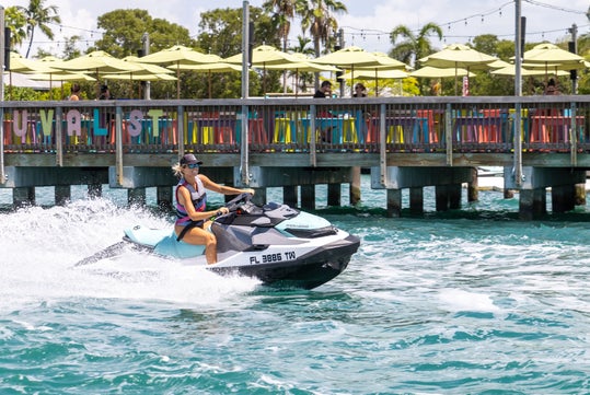 A person rides a jet ski on a sunny day near a colorful dock with people seated under green umbrellas.