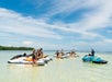 People on jet skis gather in shallow, clear water under a blue sky. A person stands on a jet ski nearby, appearing to give instructions.