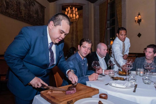 A man in a suit slices steak at a table for four seated men in a formal restaurant setting, while a server stands nearby.