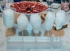 Four sticks of white cotton candy are displayed upright in a holder, with a plate of thinly sliced cured meat visible in the background.