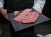 A person in formal attire holds a slate plate with several slices of marbled raw beef and a small cube of fat.