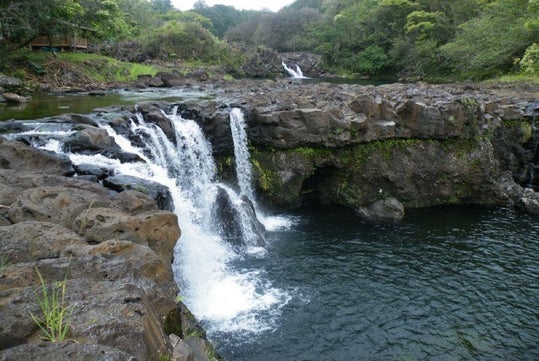 A small waterfall flows over dark volcanic rocks into a clear pool surrounded by lush green vegetation and trees.