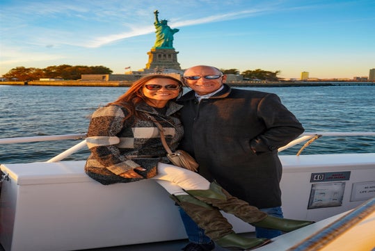 A man and woman smile while posing together on a boat with the Statue of Liberty and Ellis Island in the background on a clear day.
