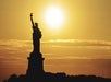 Silhouette of the Statue of Liberty against a yellow-orange sunset sky, with the sun directly behind the statue.