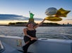 A woman in a black dress holds gold and silver balloons on a boat with the Statue of Liberty and sunset sky in the background.