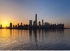 Sunrise behind the silhouette of a city skyline with tall buildings reflected in calm water.