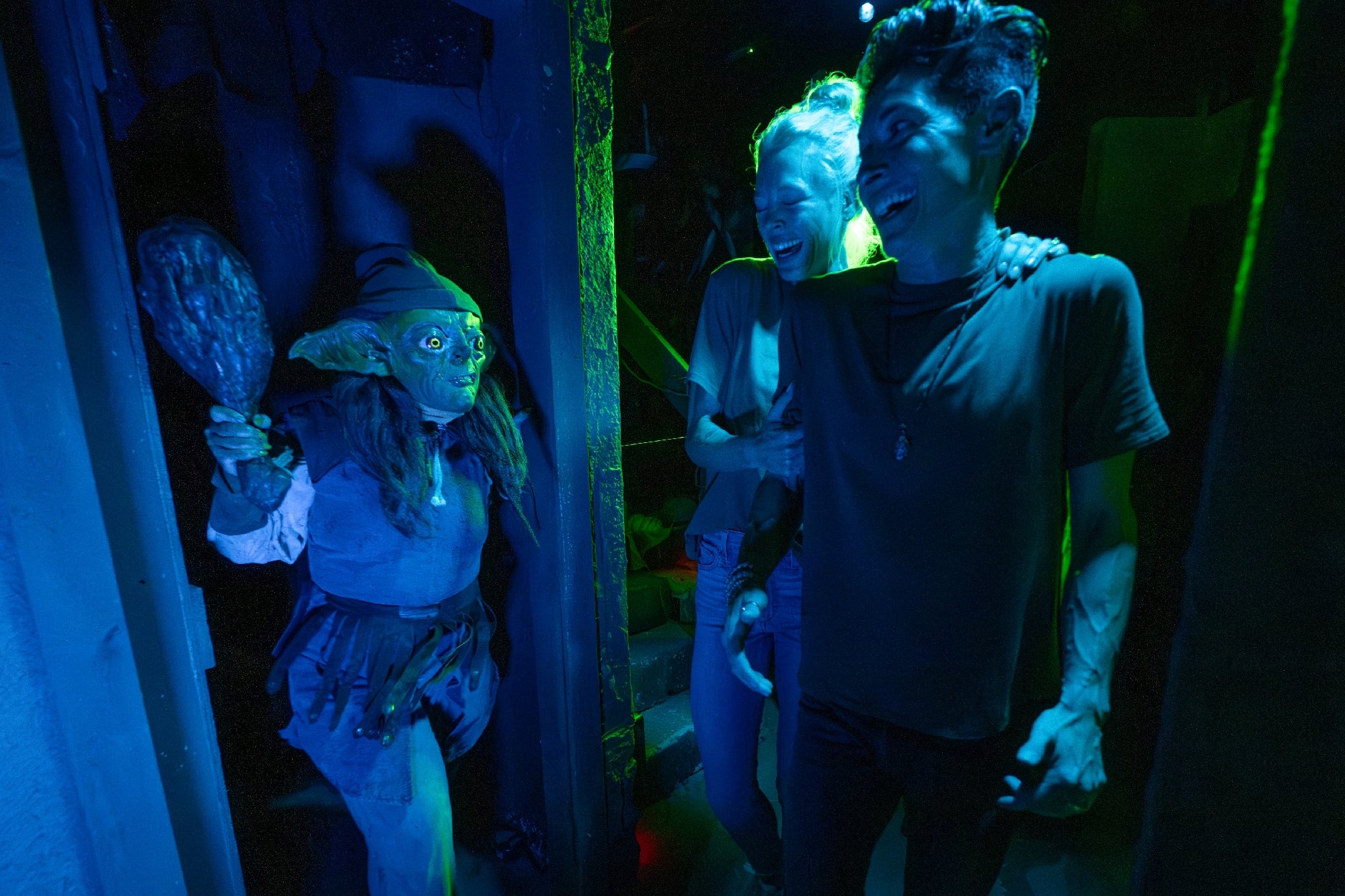 A person in a goblin costume holding a club approaches two people who appear startled, in a dimly lit, themed setting.