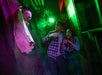Two people look startled as they encounter a scary figure emerging from a wall in a dark, foggy, green-lit hallway.