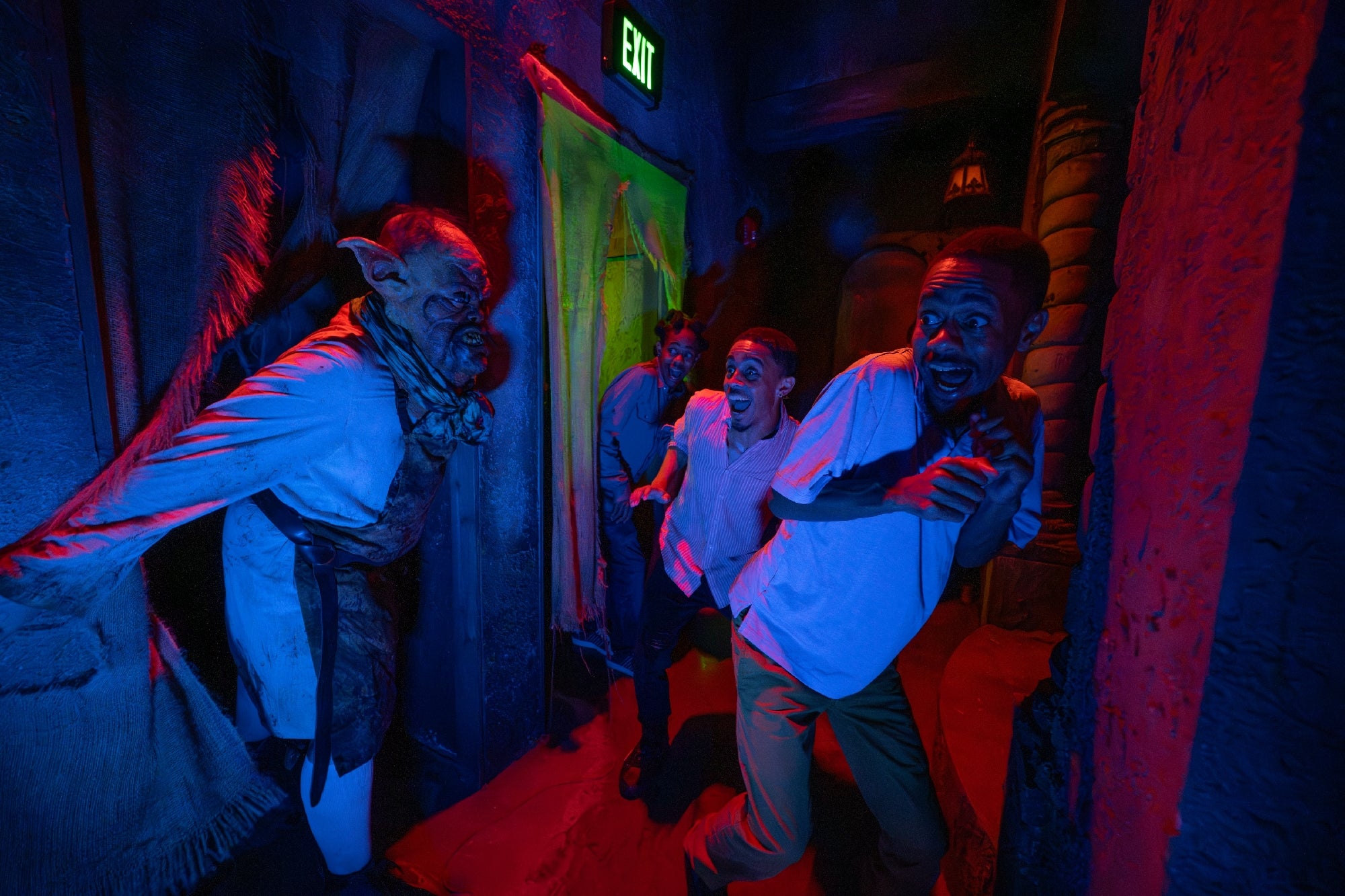 Three people react in fear as they encounter a costumed monster in a dimly lit, themed haunted house with colored lighting and an illuminated exit sign above a doorway.