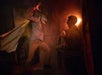 A menacing figure in a hat and coat lunges toward two startled people in a dimly lit, smoky room.