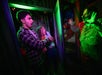 Two people react with fear as a person dressed as a scary clown approaches them in a dimly lit, neon-colored room.