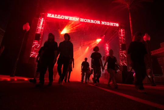 People walk under a glowing red entrance sign that reads "Halloween Horror Nights" at night, with smoke and dramatic lighting in the background.