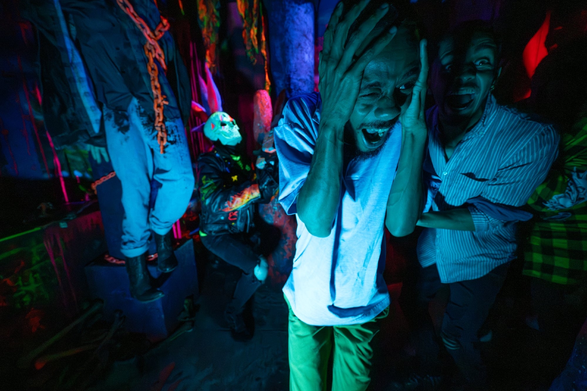 A man screams and covers his ears in a dark, neon-lit haunted house scene, with costumed figures and eerie decorations surrounding him.