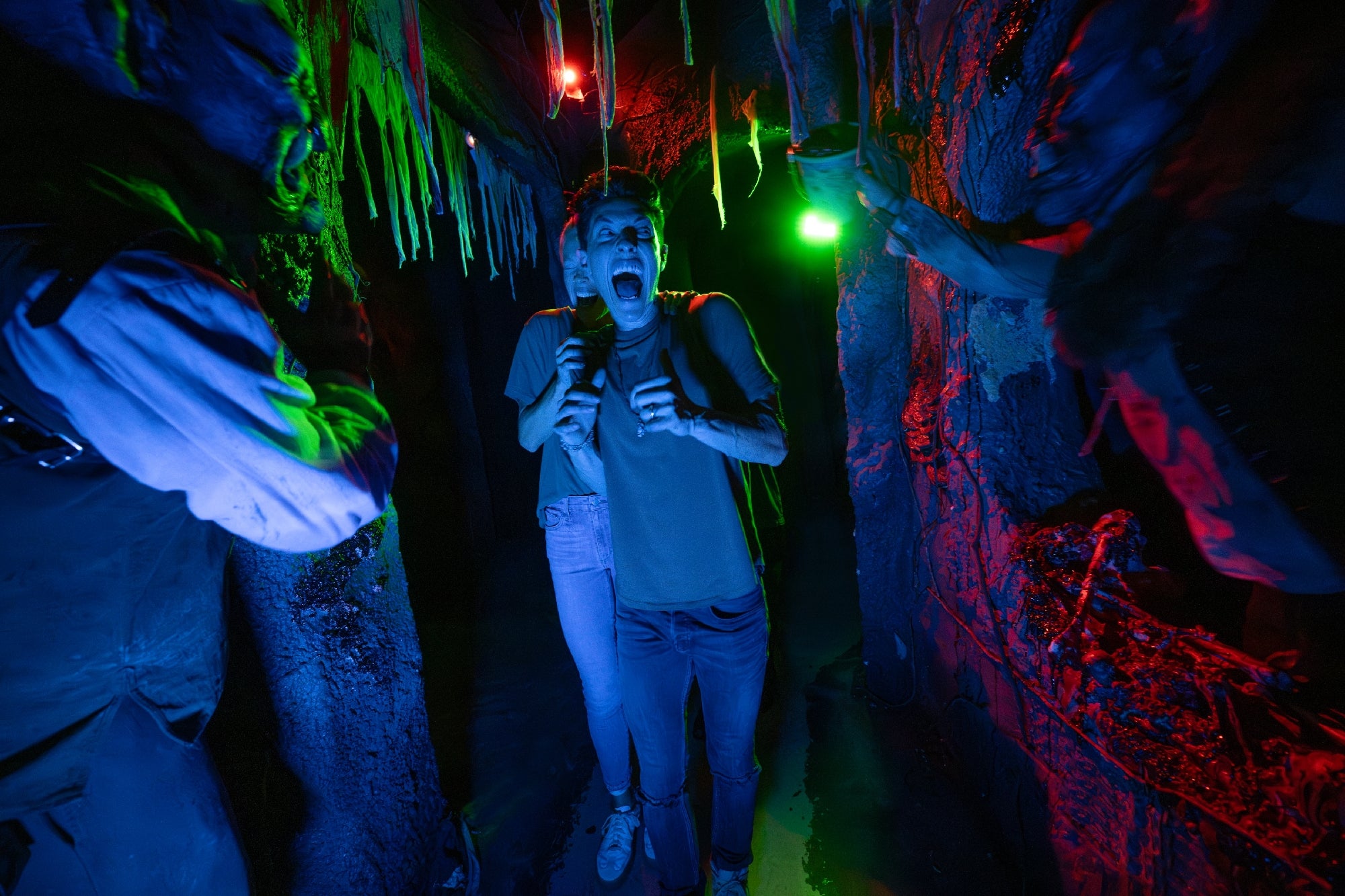 A frightened person walks through a dimly lit, spooky hallway with eerie decorations and actors in costumes reaching toward them.