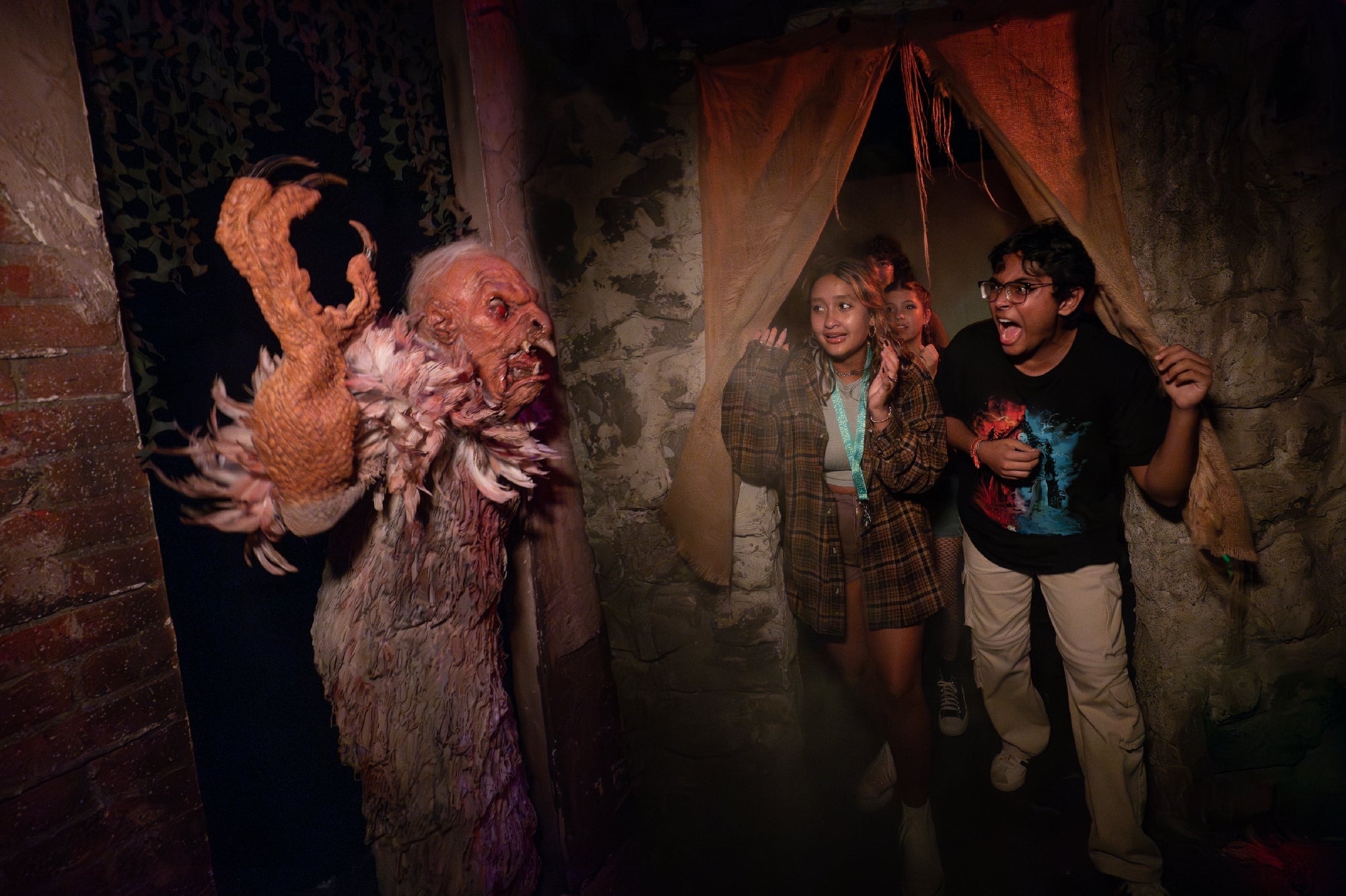 Three people react with surprise and fear as a person in a monster costume lunges at them in a dimly lit, haunted house setting.