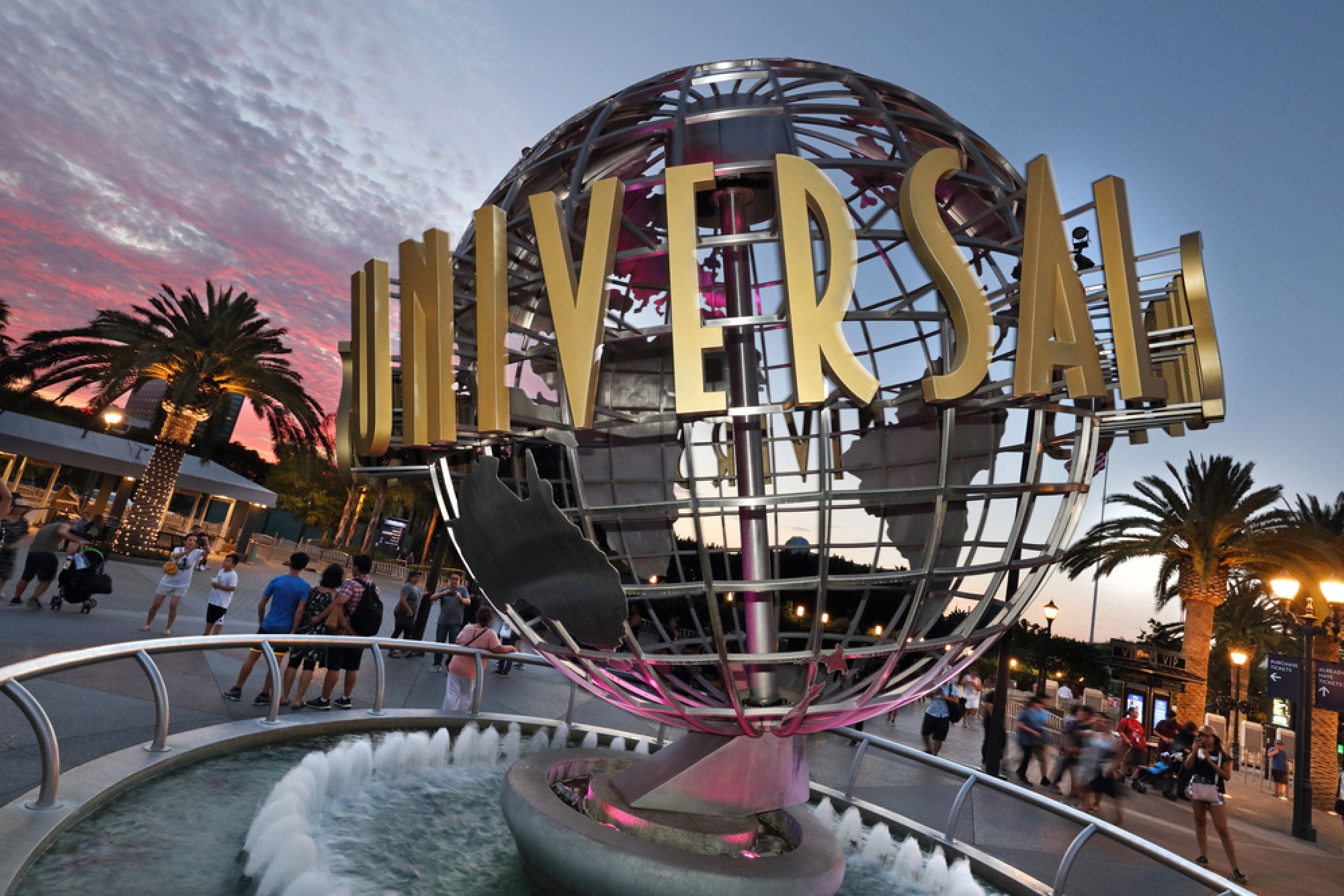 Large globe sculpture with "UNIVERSAL" text at Universal Studios entrance, surrounded by palm trees, water fountain, and people during sunset.