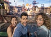 Three people take a selfie and smile excitedly in front of an outdoor water stunt show with fiery explosions and industrial set structures in the background.