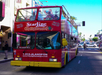 A red double-decker CitySightseeing tour bus drives down a sunny city street with people on the open top deck, passing by palm trees and shops.