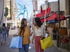 Two women holding shopping bags walk and talk on a lively urban street with a Bubba Gump restaurant and a large blue gorilla sign in the background.