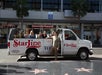 A tour van labeled "StarLine Tours" is parked on a street. People are sitting inside, and a person is standing outside, leaning on the vehicle. There are palm trees and shops in the background.