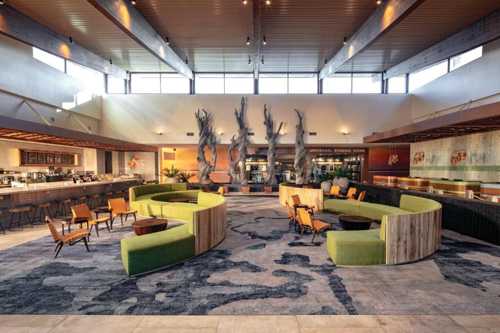 Spacious hotel lobby with modern curved green seating, wooden tables, a bar on the left, large windows, and decorative tree-like sculptures in the center.