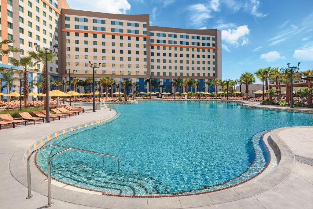 Large outdoor swimming pool surrounded by lounge chairs, umbrellas, and palm trees, with a modern multi-story hotel building in the background under a blue sky.