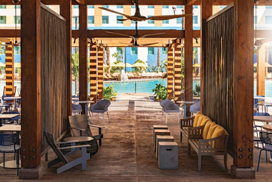 Wooden outdoor lounge area with chairs and sofas facing a swimming pool, surrounded by tables and chairs, and a building visible in the background.