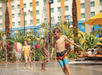 Children play in a splash pad with water jets, palm trees, and a colorful hotel building in the background. One child chases a beach ball.