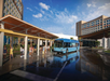 A blue shuttle bus stops outside a large hotel entrance with people waiting under a covered area on a sunny day.