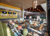 Modern hotel lobby with colorful seating, bar area in the background, large windows letting in natural light, and contemporary lighting fixtures overhead.