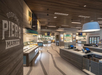Spacious, modern cafeteria with wooden decor, food display counters, self-service kiosks, and refrigerators labeled "Grab-N-Go." Sign reads "Dockside Piers Market.