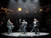 Three men in black suits sitting on stools on stage at VEGAS! THE SHOW with a band above them and lights behind them.