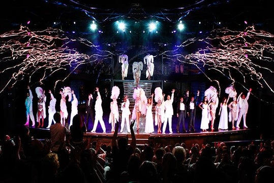 The cast of VEGAS! THE SHOW on stage in all white with streamers shooting off over their heads.
