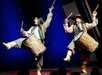 Two men in colorful vests on stage playing large drums that are strapped to their sides at V - The Ultimate Variety Show in Las Vegas, NV.