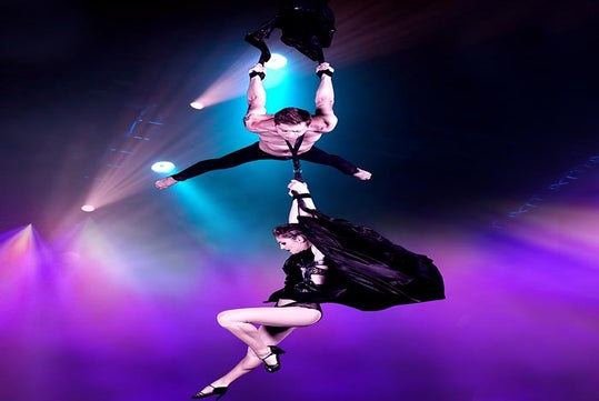 A shirtless man wearing a tie hanging from an arial silk with a woman hanging from his tie at V - The Ultimate Variety Show.