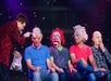 Four people wearing deformed masks sitting on stage with a man in red jacket holding a microphone to their left at V - The Ultimate Variety Show.