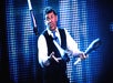 Man in a suit and tie juggling three clubs on stage with a blue dotted light background, looking up at the clubs in mid-air.
