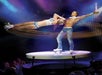 A shirtless man on roller skates with a woman also in roller skates hanging in the air from a tie around his neck in the V - The Ultimate Variety Show.