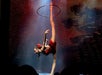 A performer in a red costume balances on one leg with the other leg extended vertically, holding a hoop above her head on a stage with a lit, dotted background.