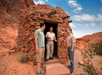 Three men stand outside a small, round stone structure in a desert landscape with red rock formations. One man is wearing a park ranger uniform.