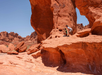 Person kneeling on reddish rocky terrain under large rock arch in a desert landscape with clear blue sky.