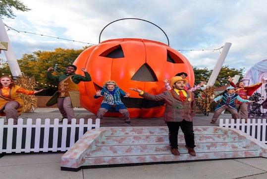 A group of performers in colorful costumes stand on a stage in front of a large jack-o'-lantern decoration, with a white picket fence and autumn trees in the background.