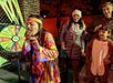 A girl in a colorful costume reacts after spinning a prize wheel, while three people in coats and costumes watch nearby at a nighttime outdoor event.